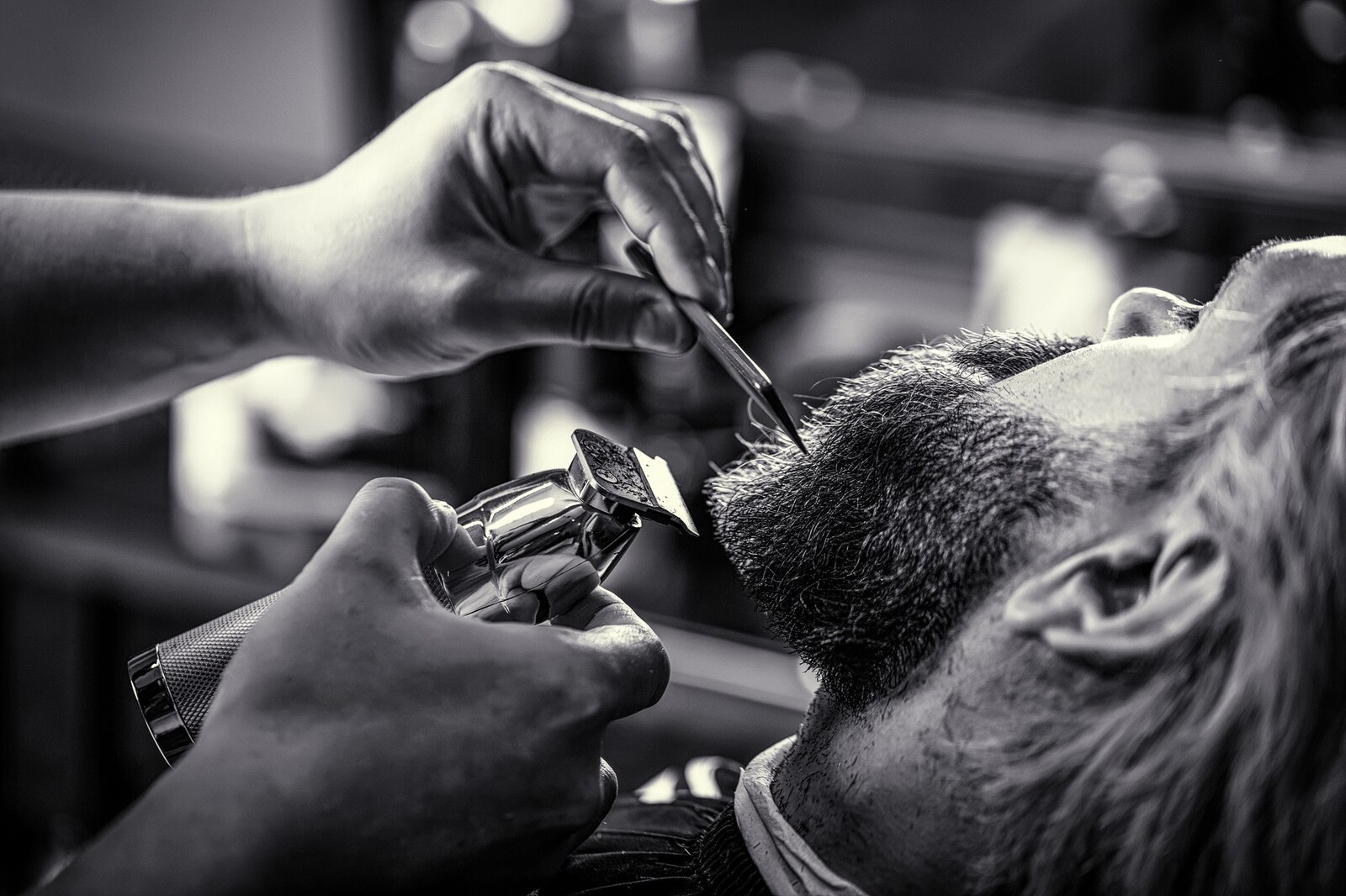 Black and white close-up of a barber's hands trimming a client's beard with a clipper.