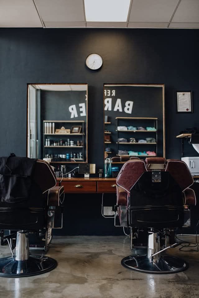 Two barber chairs at Barbier facing framed mirrors on a dark wall, with shelves of bottles and folded towels reflected behind them.