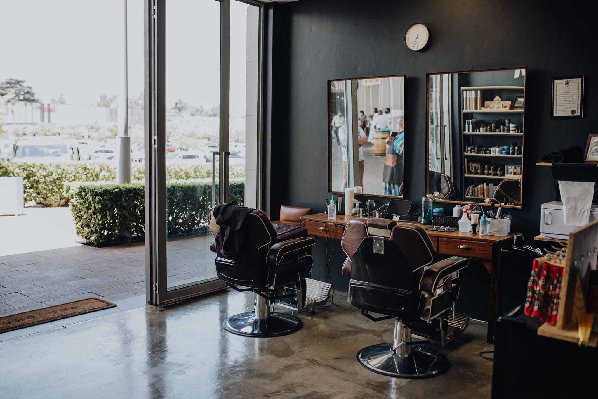 Inside Barbier: two leather barber chairs facing framed mirrors on a dark wall, a counter of tools and bottles beside them, and the glass shopfront standing open to the street.