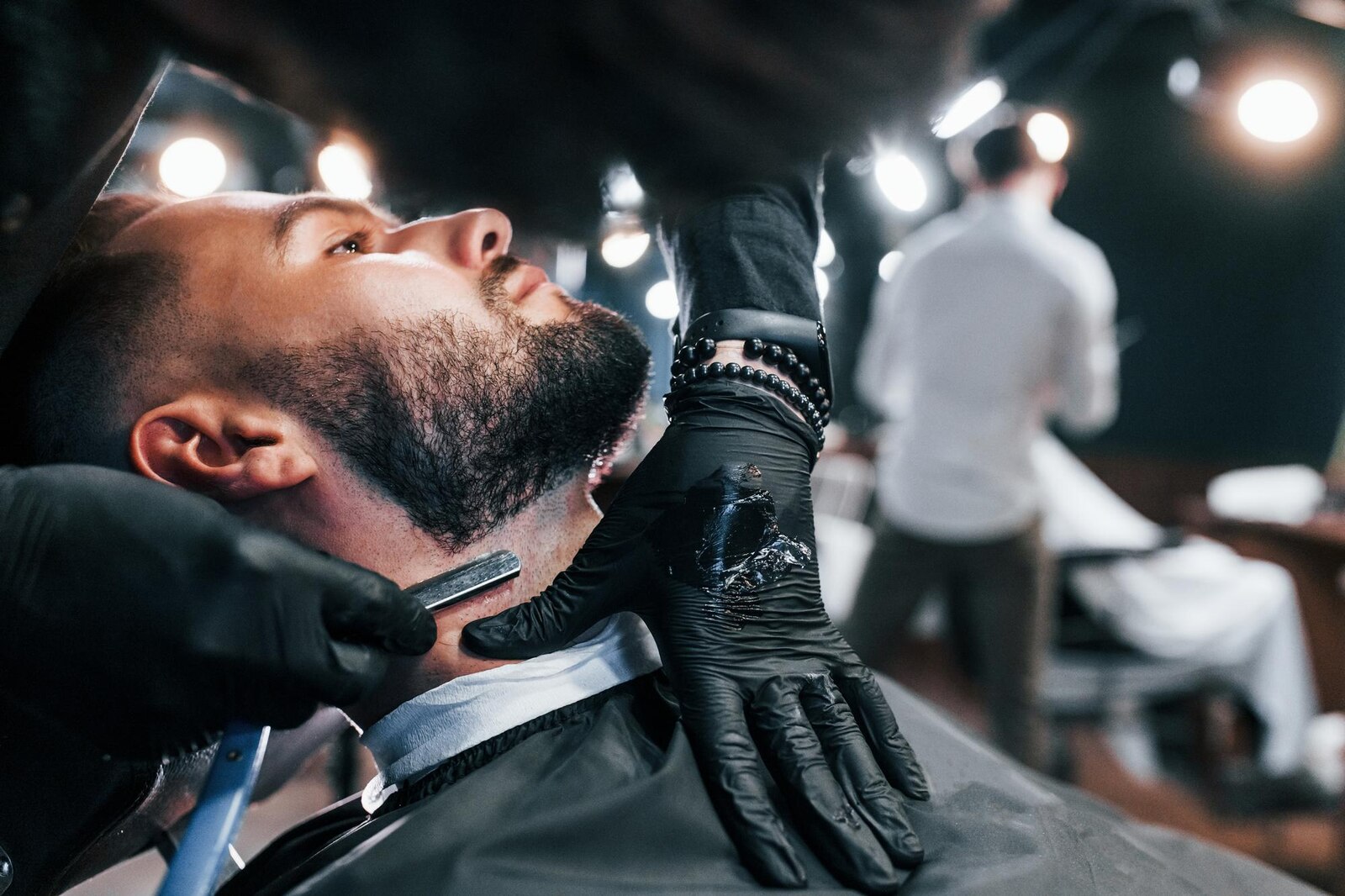 A young man with a styled haircut sitting in a barber shop chair while his beard is shaved.